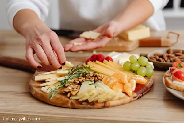 woman putting together a charcuterie board of cheeses, fruits, cracker, nuts